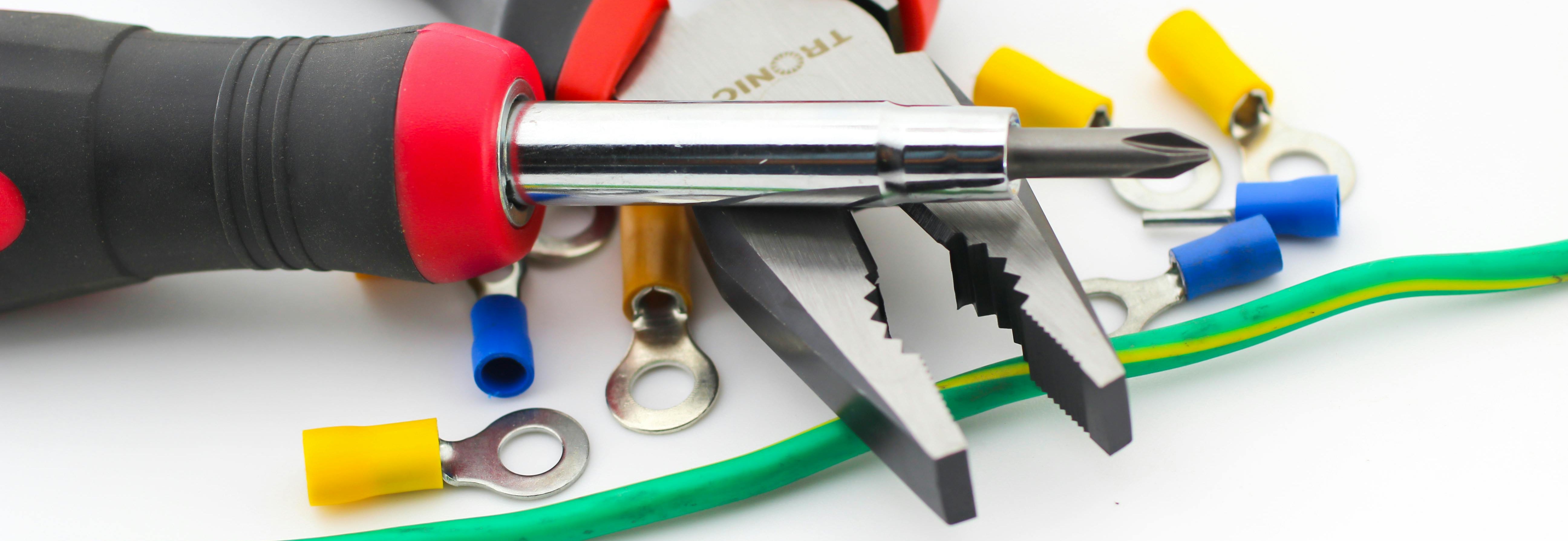 Close-up of red-and-black handled pliers and a screwdriver surrounded by colored crimp ring terminals and a green insulated wire on a white background.